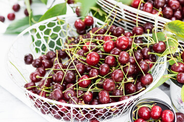 Freshly gathered juicy red cherries  in white metal containers closeup , red cherries in garden on white wooden table in cherry tree leaves background