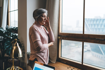 Senior woman standing near window having migraine