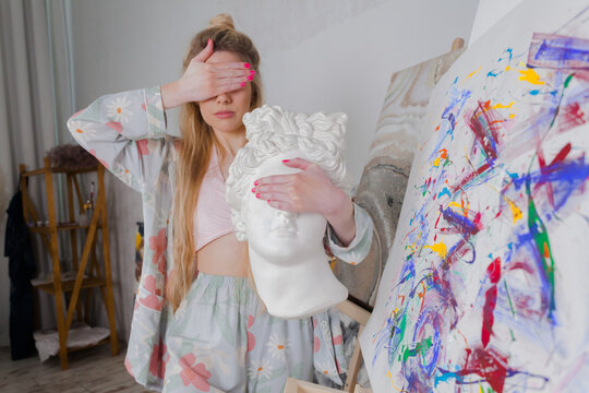 Talented Female Artist smiling, holding marble head and covering her eyes. Colorful, Emotional, Sensual Painter Creating Abstract Modern Art. Creative Expression concept.Paintings in the background.