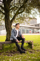 Obraz premium young latin man sitting resting on a park bench in the shade of a leafy tree, relaxed lifestyle surrounded by nature, portrait