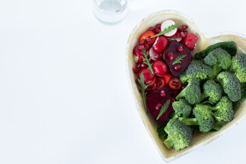 Colorful plant-based food (fruits and vegetables) in heart-shaped plate with glass of fresh water isolated on white background. Detox, healthy eating, anti-cancer diet concept. Top view. Copy space.
