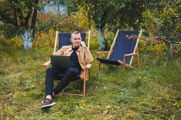 A handsome man in a brown shirt sits on a wooden folding chair in the garden and works with a laptop. Wooden ecological furniture. Remote work