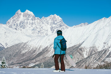 Female Snowboarder standing back to camera out of focus with snowboard looking to beautiful mountain peaks covered with snow