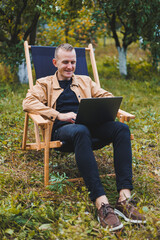 Smiling young freelancer man sitting in wooden chair with laptop while working outdoors in garden. Remote work.