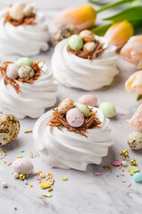 Fototapeta na wymiar Easter treat - set of white meringues in shape of nest with multicolored candy chocolate eggs, tulips and sprinkles over marble background. Side view, close up. Holiday symbol