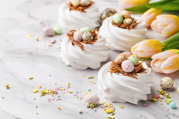 Easter treat - set of white meringues in shape of nest with multicolored candy chocolate eggs, tulips and sprinkles over marble background. Side view, close up, copy space. Holiday symbol