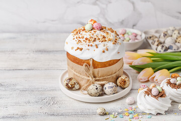 Traditional easter cake or sweet bread, quail eggs, candy chocolate eggs, tulips and white meringues in shape of nest over white wooden table. Side view, copy space. Easter treat, holiday symbol