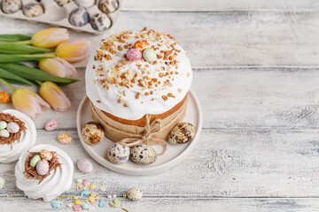 Traditional easter cake or sweet bread, quail eggs, candy chocolate eggs, tulips and white meringues in shape of nest over white wooden table. Top view, copy space. Easter treat, holiday symbol