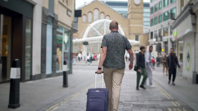 Male tourist walks pulling suitcase through London city streets, looks at phone