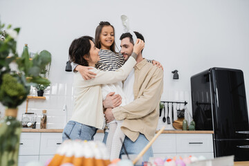 Smiling girl hugging parents near blurred Easter eggs in kitchen.