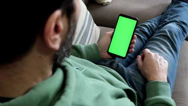 Man sitting on sofa makes single finger tap on smartphone green screen.