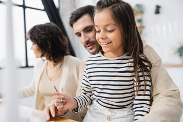 Smiling girl holding paintbrush near blurred parents at home.