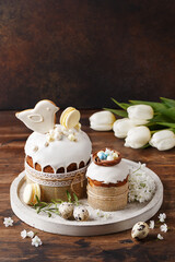 Set of traditional easter cakes with icing, decorated with meringue and gingerbread cookie in shape of bird over dark brown background with tulips. Side view, close up. Easter treat, holiday symbol