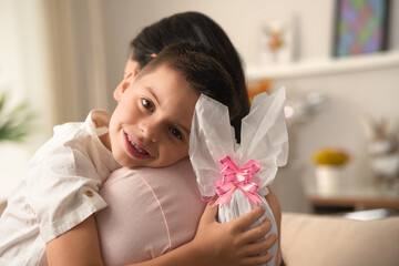 Joyful brazilian child looking at camera and hugging mother with easter egg in the hand at home