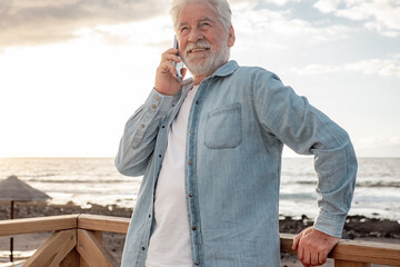 Portrait of beautiful senior bearded man in casual shirt standing outdoors at beach in sunset light using mobile phone. Smiling old man talking on smartphone