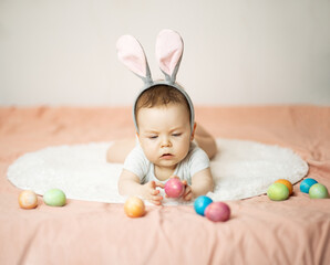 A focused infant baby is lying on the bed, wearing bunny ears and holding a color Easter egg in his hands. Hunt on the Easter morning. Copy space.