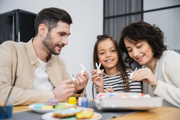Obraz premium Smiling kid holding Easter rabbit figurine near parents and eggs at home.