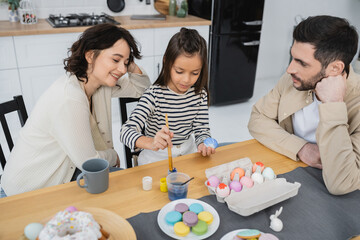 Smiling woman sitting near daughter coloring Easter eggs and husband at home.