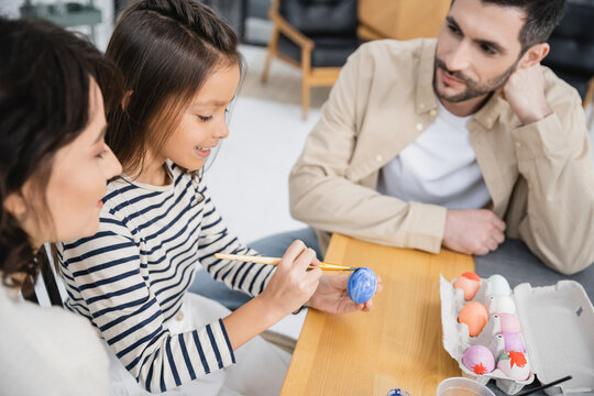 Smiling girl painting Easter egg near mother and father at home.