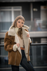 Gorgeous smiling girl in stylish clothes posing on modern balcony with amazing view on city .Portrait of a Stylish fashionable blonde  teenage with fur coat with large collars wearing in balcony