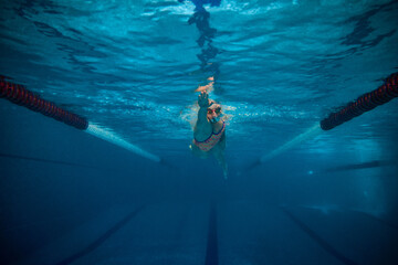 Fast and strong. Professional female swimmer in cap and goggles in motion, training in swimming pool indoor. Underwater view. Concept of sport, endurance, competition, energy, healthy lifestyle