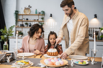 Father cutting Easter cake near daughter and wife during festive dinner.