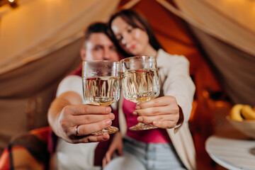Happy lovely couple relaxing in glamping on summer evening and drinking wine near cozy bonfire. Luxury camping tent for outdoor recreation and recreation. Lifestyle concept