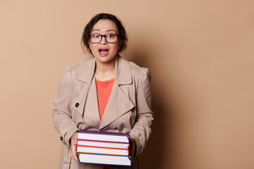 Astonished multi-ethnic woman wearing beige coat and eyeglasses, a school teacher holding heavy volumes of hardcover books, expressing stupefaction looking at camera, isolated over cream background