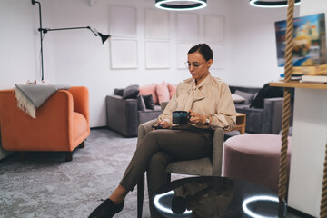 Focused female using smartphone and drinking hot beverage in office