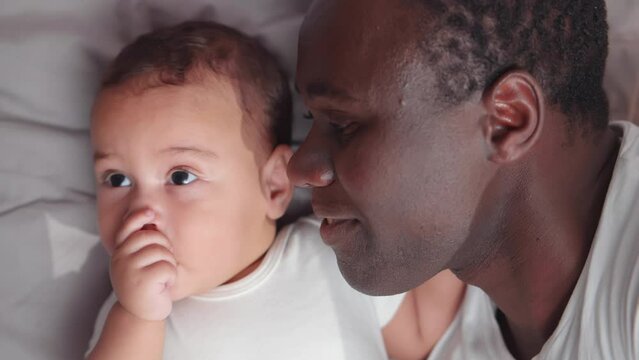 Adorable African American baby lying with father in bed. Realtime