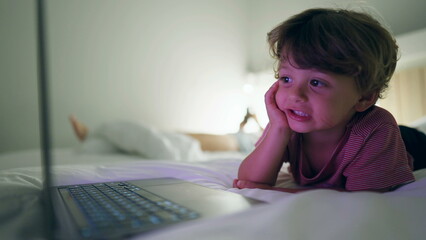 Young boy starring at media entertainment on laptop screen laying in bed at night. Hypnotized kid staring at blue glowing screen in the evening