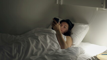 Woman looking at cellphone device laying in bed at night. Female person staring at glowing screen in the dark before bedtime