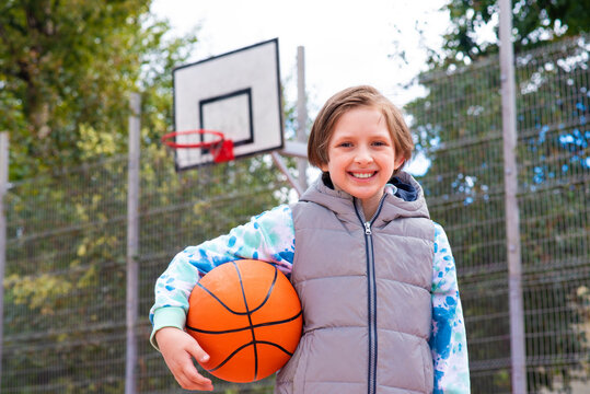 a sports boy in a sweater and vest stands on the basketball court, holds the ball in his hands, spring or autumn outside