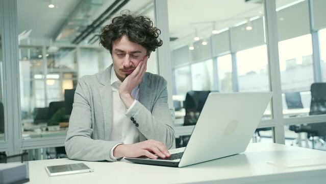 Sad young business man freelancer with severe toothache suffers from strong pain ache sitting at desktop in workplace office Unhealthy entrepreneur bothered by sore tooth while typing on laptop indoor