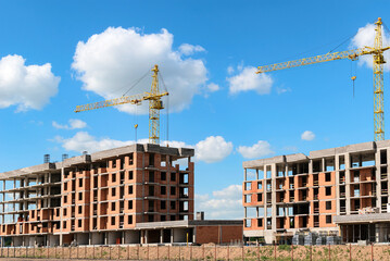 Construction site with multi-storey residential buildings under construction and cranes against a blue sky with clouds.