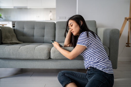 Downcast asian woman looking sadly to device screen reading unpleasant message or waiting for call. Upset Chinese girl using cellphone grudgingly reading news pondering problem considering solutions.