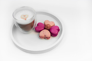 Cappuccino with a heart on the foam and heart-shaped macarons laid out like a flower on a white stand, white background. St. Valentine's Day romantic breakfast. Sweets with coffee.