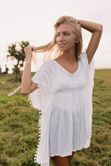 Romantic adorable stylish woman with long blond hair and charming smile posing during photoshoot on the shore of ocean in sunlight in summer day. 