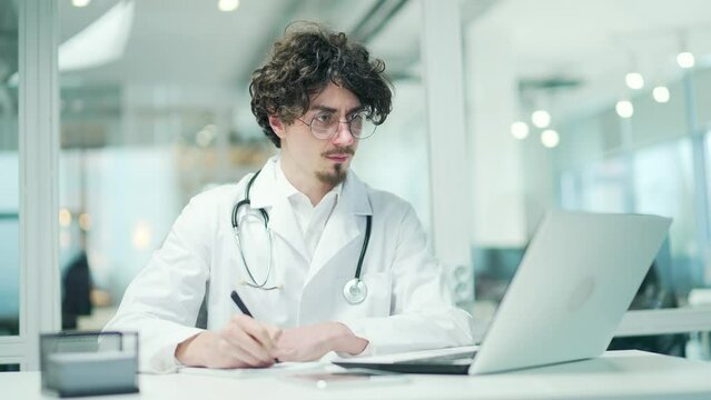Focused young doctor therapist with stethoscope looking at laptop screen and writing treatment guidelines for patient or studying at hospital office indoors Distance remote medical education concept