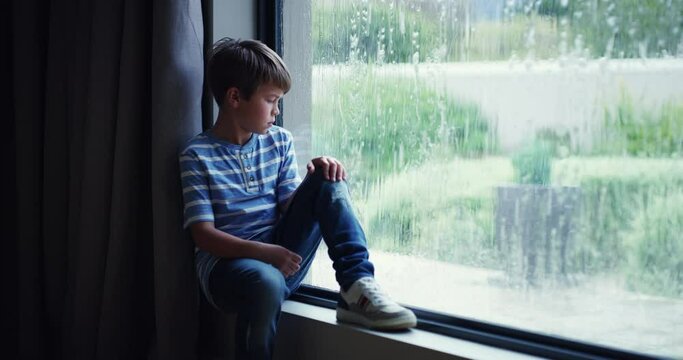Sad, depressed and frustrated child with mental health problems watching the rain through a window at home. Abused, unhappy little boy looking lonely and upset, thinking of unhappy news of divorce