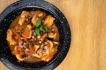 Pasta. Top view of traditional stuffed agnolottis with a tomato sauce with mushrooms, bacon and basil in a black dish on the wooden table.