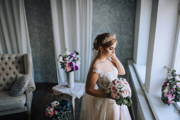 A beautiful young blonde bride in a white lace dress with a crown, a diadem on her head stands in a studio, indoors with a bouquet in her hands near the window. Wedding photography, portrait.