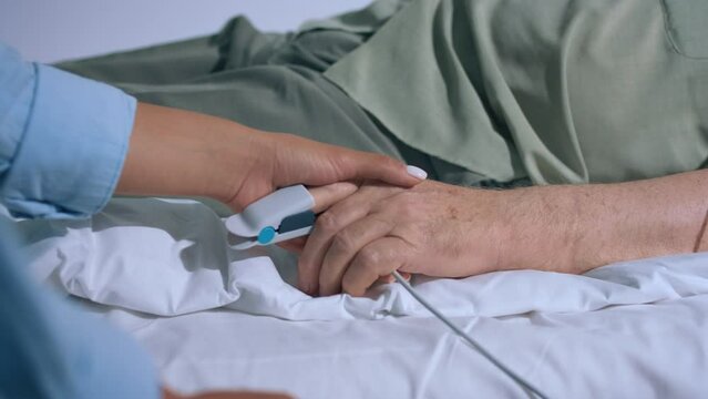 Closeup to the camera old hand patient wearing the finger heart rate and young hand holding and supporting the patient in the hospital room video details capturing