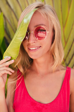 Close-up portrait of smiling woman on rest in pink t-shirt and round glasses. Curly blond girl with snow-white smile looks into camera on background of tropical plants