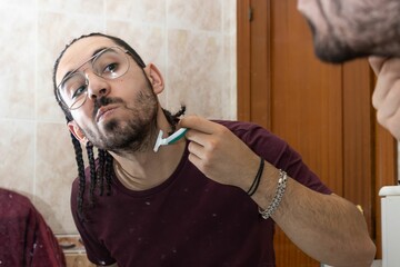 Man shaving his beard with a razor in front of the mirror. Reflection of young man shaving his beard in the mirror. Adult man shaving his beard with a razor