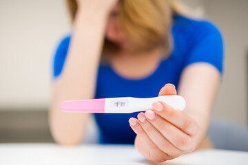 Single sad woman holding a positive pregnancy test in the living room at home.