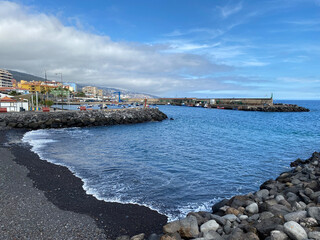 Felsige schwarze Bucht, rechts liegen Steine, links ein buntes Dorf. Blaues Meer draußen ein Wellenbrecher. Blauer Himmel mit teils mit weißen Wolken bedekt..