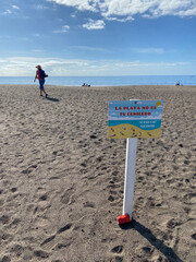 "Der Strand ist kein Aschenbecher" steht auf einer Tafel und auf spanische im Vordergrund. Dahinter Sandstrand eine Person dann im Horizont blaues Meer.