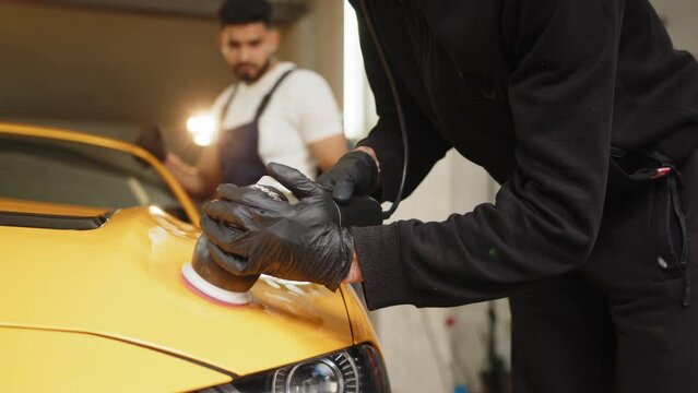 Car detailing and polishing concept. Hands of professional car service male worker, with orbital polisher, polishing yellow luxury car hood in auto repair shop.