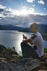 Watching cell phone sitting on rock on top of mountain at sunset in front of Patagonian Lake.